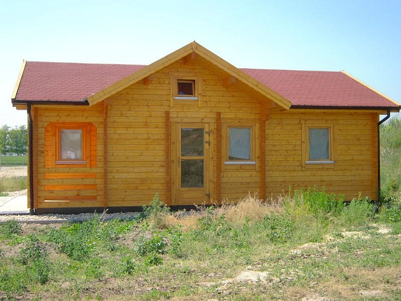 Een kleine houten hut met een rood dak, een veranda aan de voorkant en verschillende ramen, gelegen in een droog grasveld.
