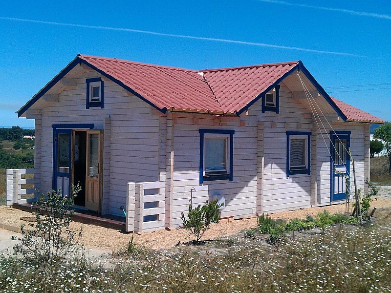 Een klein houten huis van één verdieping met een rood pannendak, blauwe luiken en een veranda aan de voorkant, gelegen in een droog grasveld onder een helderblauwe lucht.