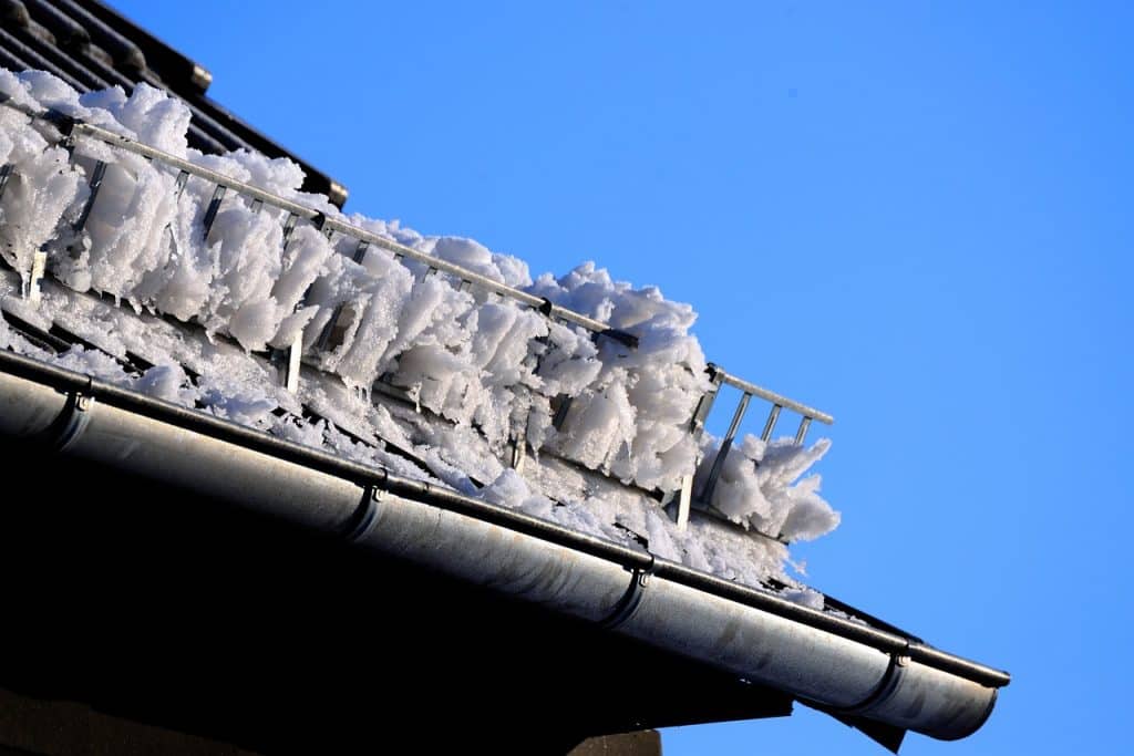 IJspegels en sneeuw hopen zich op in een dakgoot onder een helderblauwe hemel, met een kleine antenne zichtbaar boven de ijzige formaties. "Houd je houten huis droog - regengoten