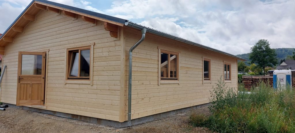 Een nieuw gebouwde houten hut van één verdieping met meerdere ramen en een enkele deur, tegen een achtergrond van heuvels en een gedeeltelijk bewolkte hemel, is met zijn houten constructie een voorbeeld van duurzaam bouwen.