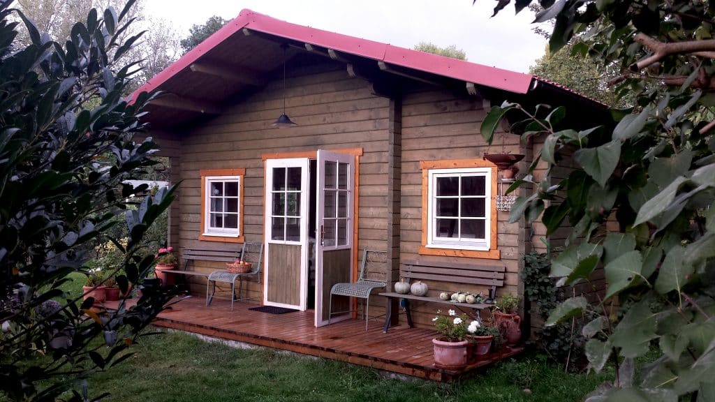 A quaint log cabin with red roofing sits surrounded by lush greenery and trees in your new garden. The cabin has open white French doors, a small porch with chairs, and several potted plants around