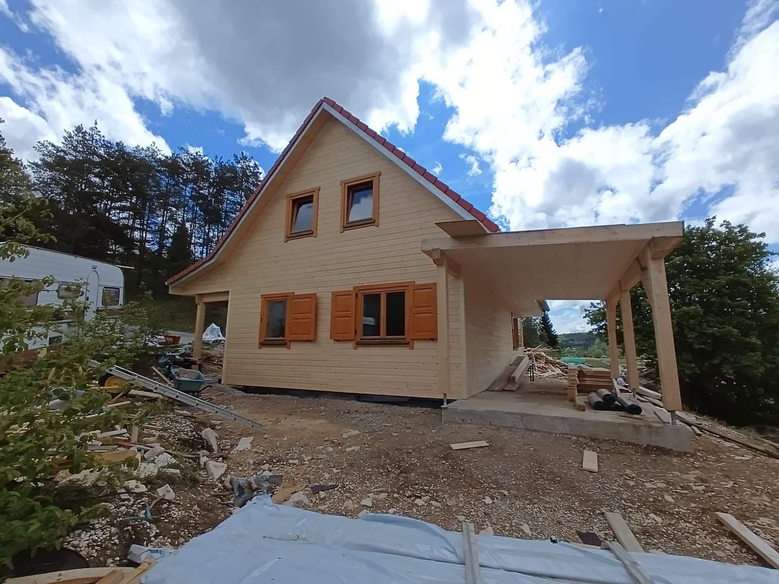 Een klein, gedeeltelijk afgebouwd houten huis met een schuin dak en rode dakspanen staat op een onverhard perceel. Het huis heeft twee ramen met bruine luiken op de bovenverdieping en twee ramen op de begane grond. Bouwmaterialen liggen verspreid over het terrein.