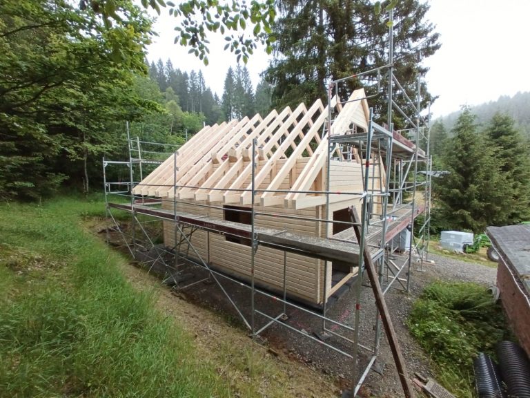A luxury wooden house under construction, surrounded by scaffolding. The roof truss is partly made of wooden beams and the walls are currently under construction. The property resembles a charming log cabin, nestled in a wooded area, with trees and green spaces in the background.