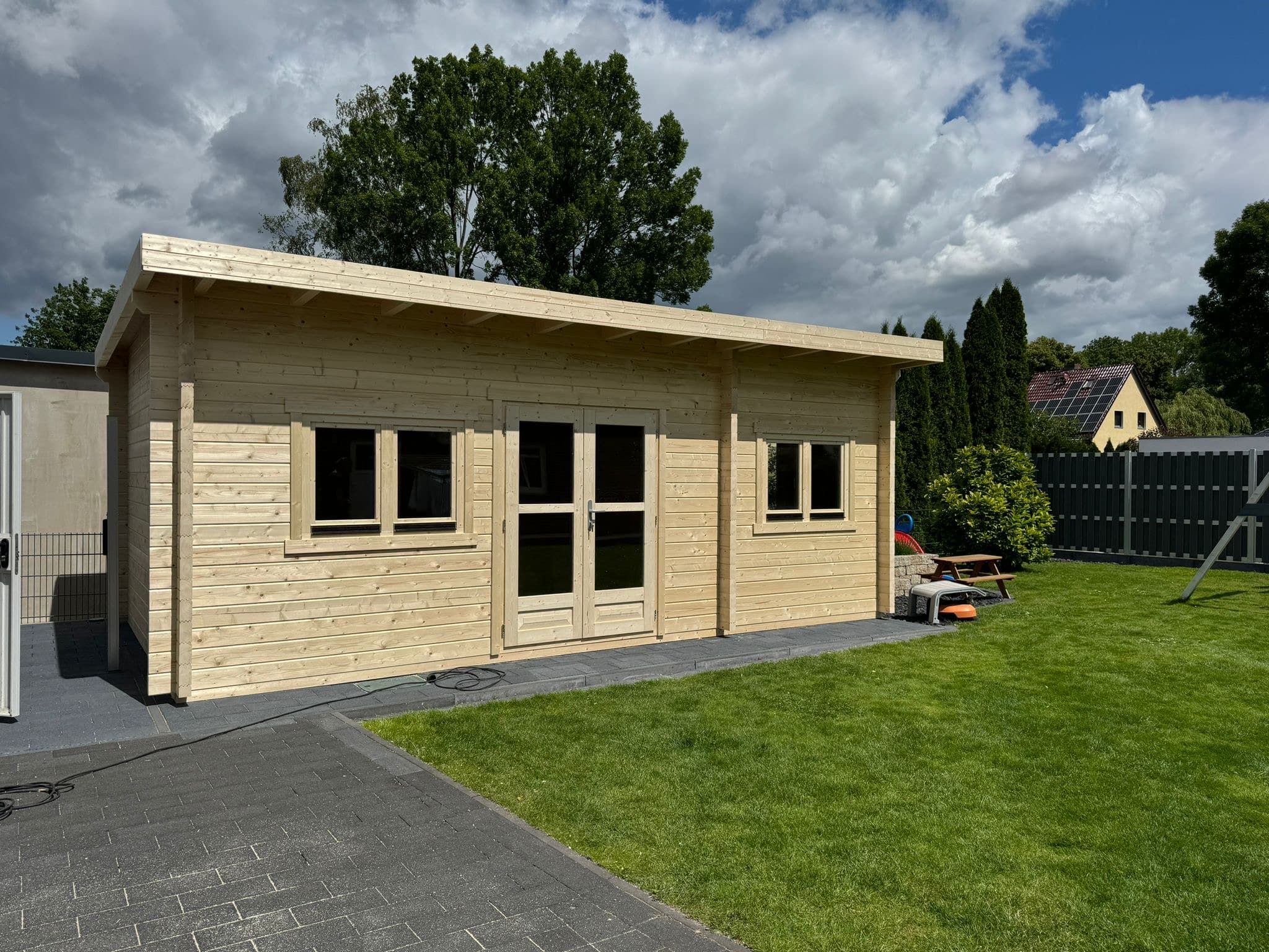 A wooden single-pitched roof house with double doors and two windows on each side stands on a paved area surrounded by a green lawn. Trees, a fence and part of a house can be seen in the background. The sky is partly cloudy. This 6500 x 3700 structure has durable, 70 mm thick walls for added stability.
